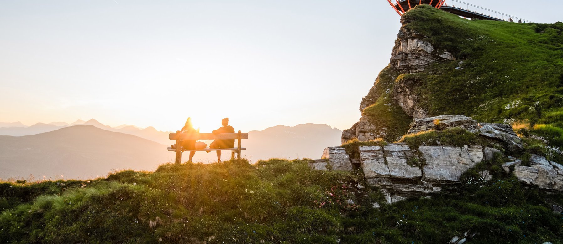 Dieses Bild zeigt die Aussichtsplattform am Stubnerkogel und eine Bank auf der zwei Menschen sich den Sonnenuntergand und das Bergpanorama anschauen.