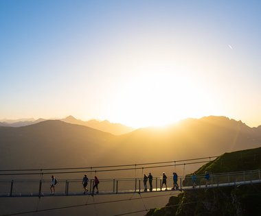 Dieses Bild zeigt eine Gruppe von Menschen, die über die Hängebrücke des Stubnerkogels während eines Sonnenunterganges gehen.