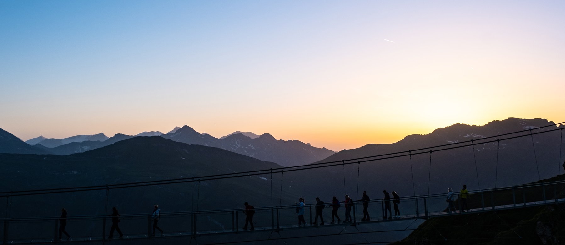Dieses Bild zeigt die Hängebrücke auf dem Stubnerkogel bei Sonnenuntergang. Eine Gruppe läuft hinüber.