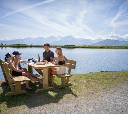 Das Bild zeigt eine Familie mit Kindern, die neben einem See auf der Schlossalm jausnet.